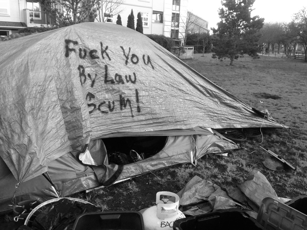 A black and white photo of a tent in a city park with a tarp over it that reads with spraypaint: "Fuck you bylaw scum".